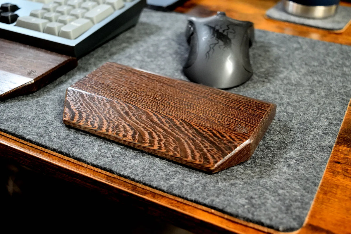 A Lonoke Designs Wenge Mouse Wrist Rest with a prominent wood grain pattern, resting on a gray wool felt desk mat on a wooden desk next to a keyboard and mouse.