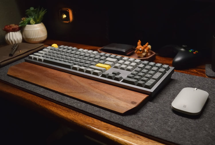 A modern desk setup featuring a gray mechanical keyboard with a walnut wrist rest placed on a dark wool felt desk pad. To the side sits a wireless mouse, a leather journal with a pen, and small accessories like a wallet and potted plants.