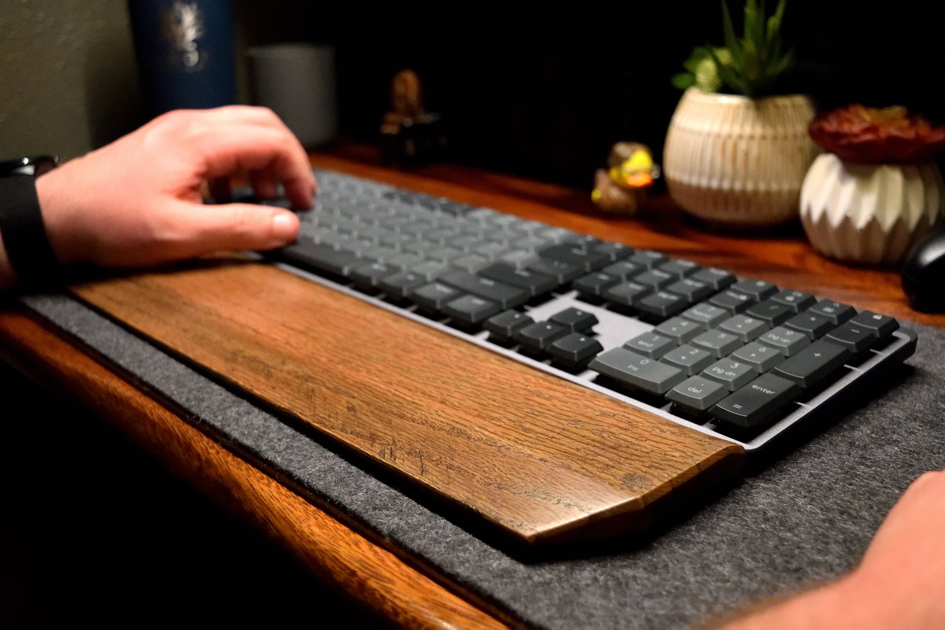 Person using a mechanical keyboard on a desk with a Lonoke Designs black-stained oak wood wrist rest, on a gray wool desk pad.