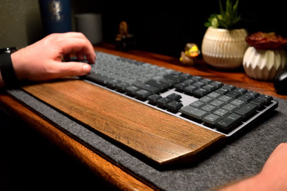 Person using a mechanical keyboard on a desk with a Lonoke Designs black-stained oak wood wrist rest, on a gray wool desk pad.