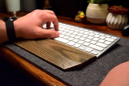Person using a wireless keyboard on a desk with a black-stained oak wood wrist rest and decorative items in the background.