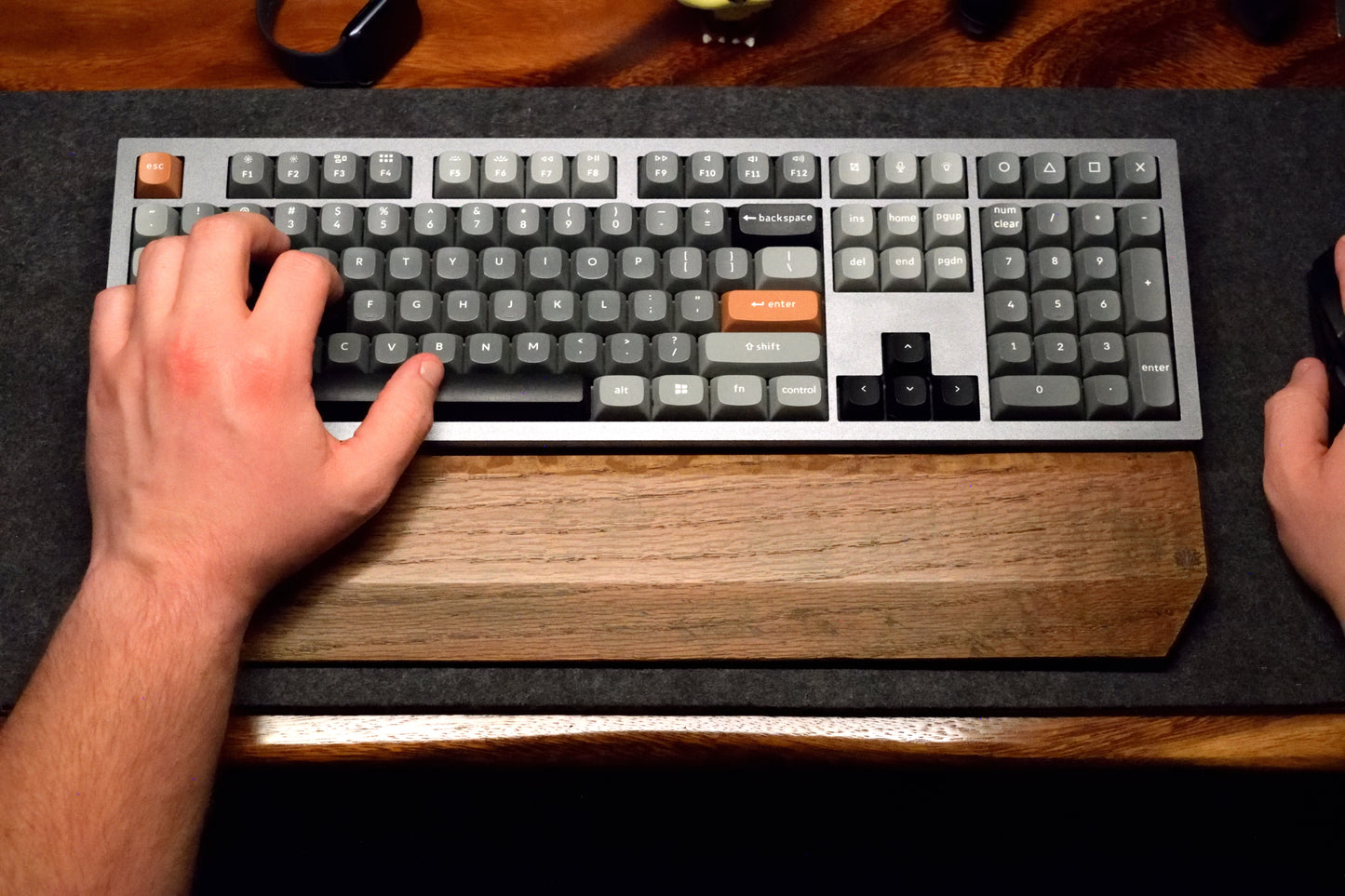 Person typing on a mechanical keyboard with a Lonoke Designs black-stained oak wrist rest on a dark wool desk pad.