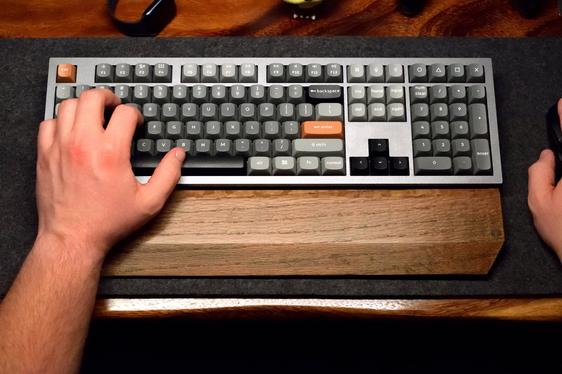 Person typing on a mechanical keyboard with a Lonoke Designs black-stained oak wrist rest on a dark wool desk pad.