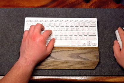 Person using a white keyboard and mouse with a black-stained Oak keyboard wrist rest by Lonoke Designs on a wool desk pad and wooden desk.