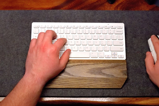 Person using a white keyboard and mouse with a black-stained Oak keyboard wrist rest by Lonoke Designs on a wool desk pad and wooden desk.