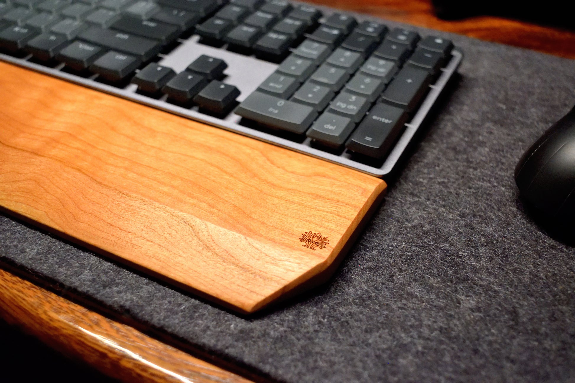 Black keyboard with a Lonoke Designs Cherry hardwood wrist rest on a wooden desk and gray wool desk pad.