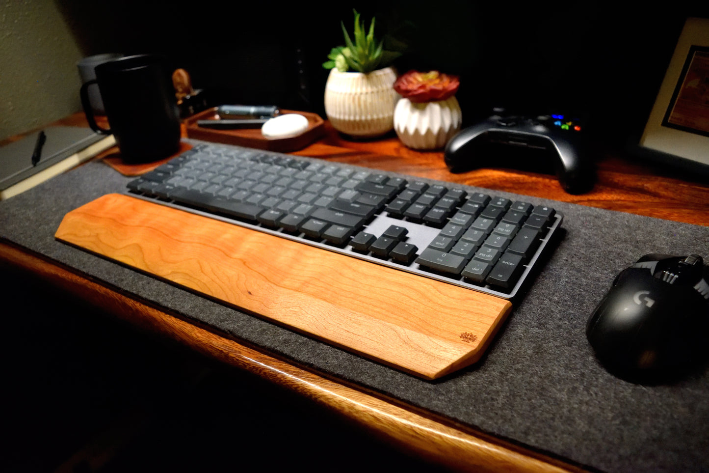Keyboard with a Lonoke Designs Cherry hardwood wrist rest on a wooden desk with a mouse, remote control, and plants in the background.