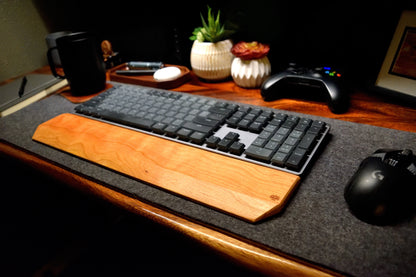 Keyboard with a Lonoke Designs Cherry hardwood wrist rest on a wooden desk with a mouse, remote control, and plants in the background.