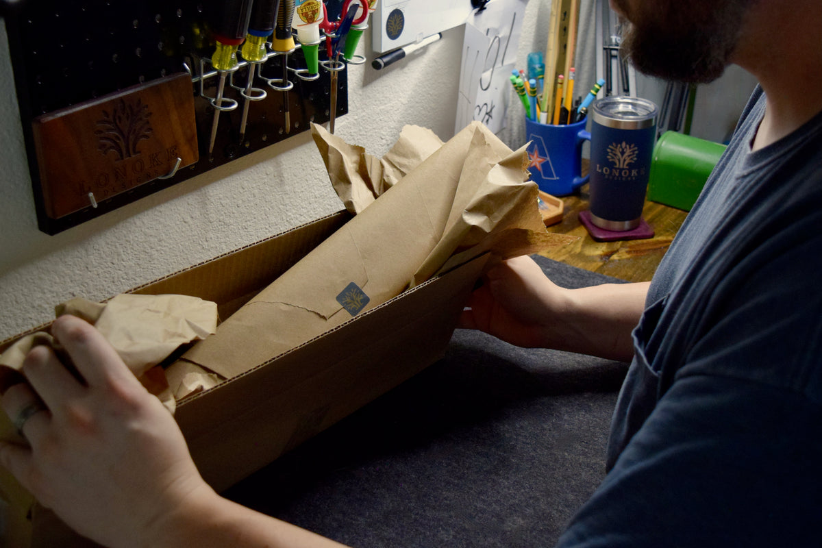 A person unpacking a long cardboard box lined with brown paper at a workbench, with a blue Lonoke Designs tumbler and desk supplies visible in the background.