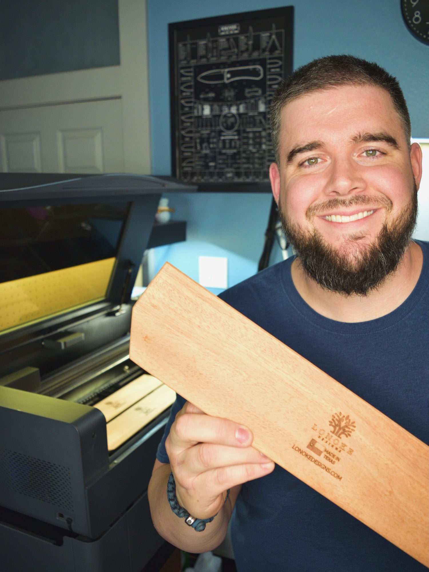 A man holding a Mahogany wrist rest with a Lonoke Designs logo, smiling in front of an engraving machine.