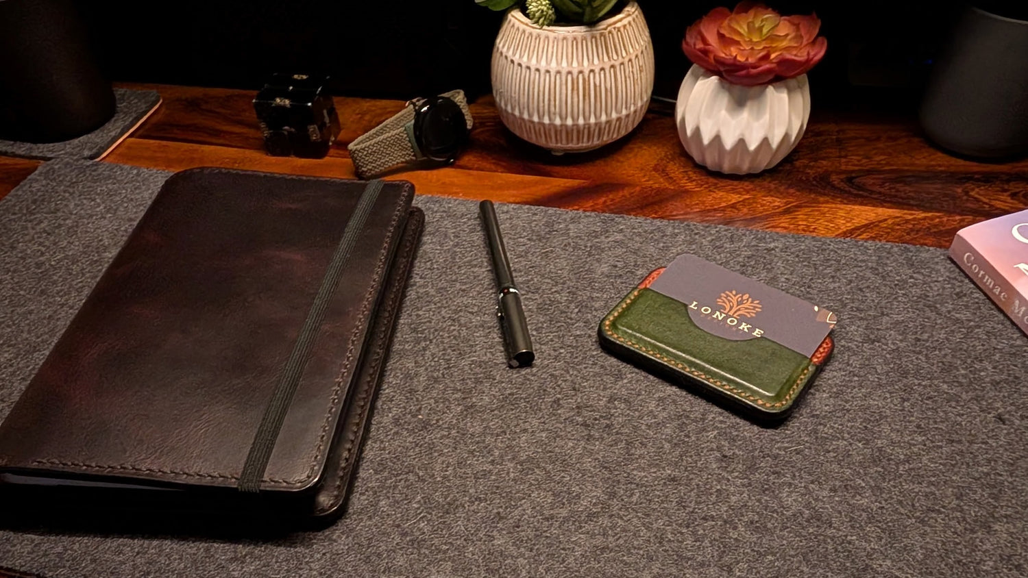 Desk setup with a leather notebook, pen, and a card with a logo on a gray surface.