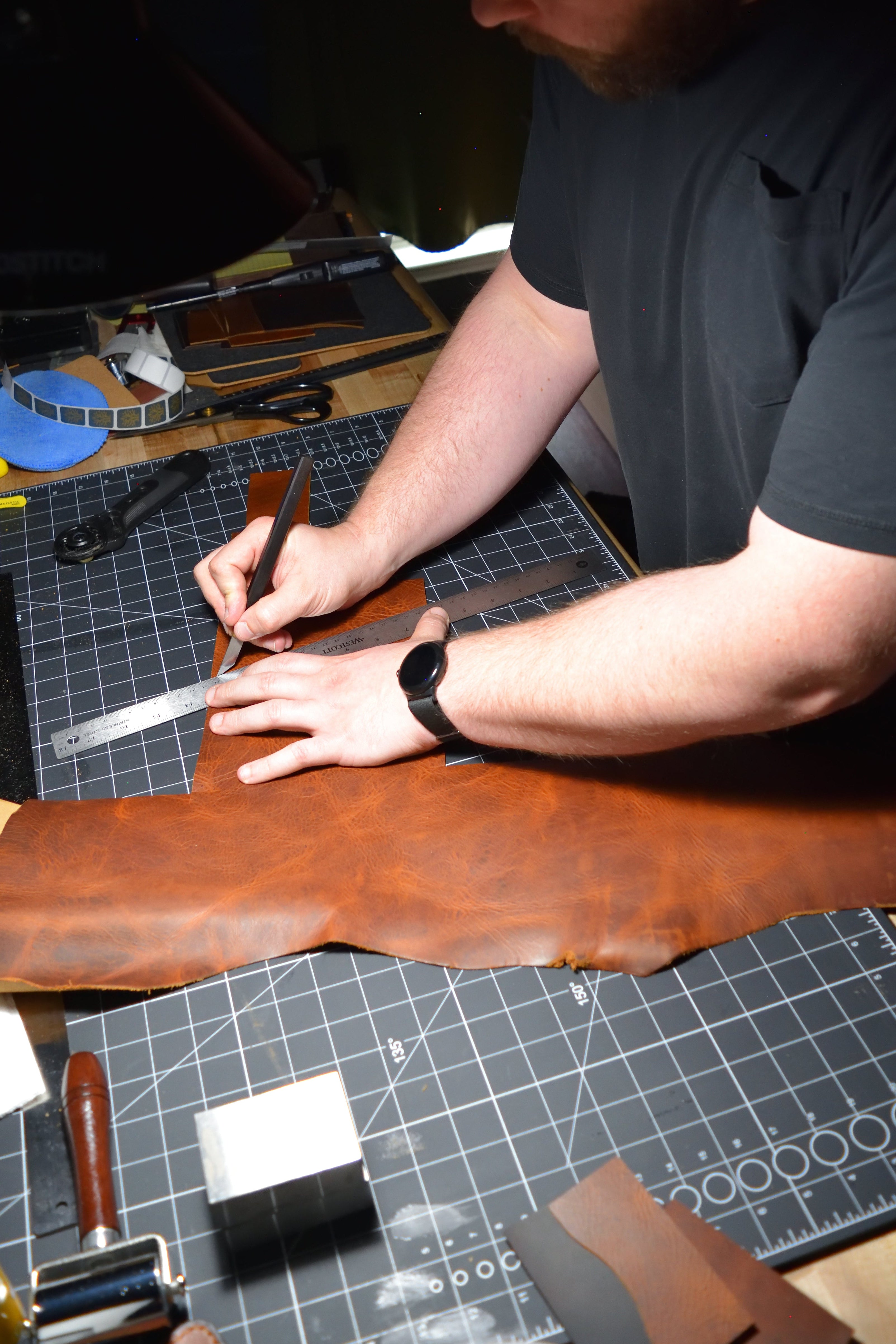 A man working with leather at a workbench with leather and tools at the Lonoke Designs workshop