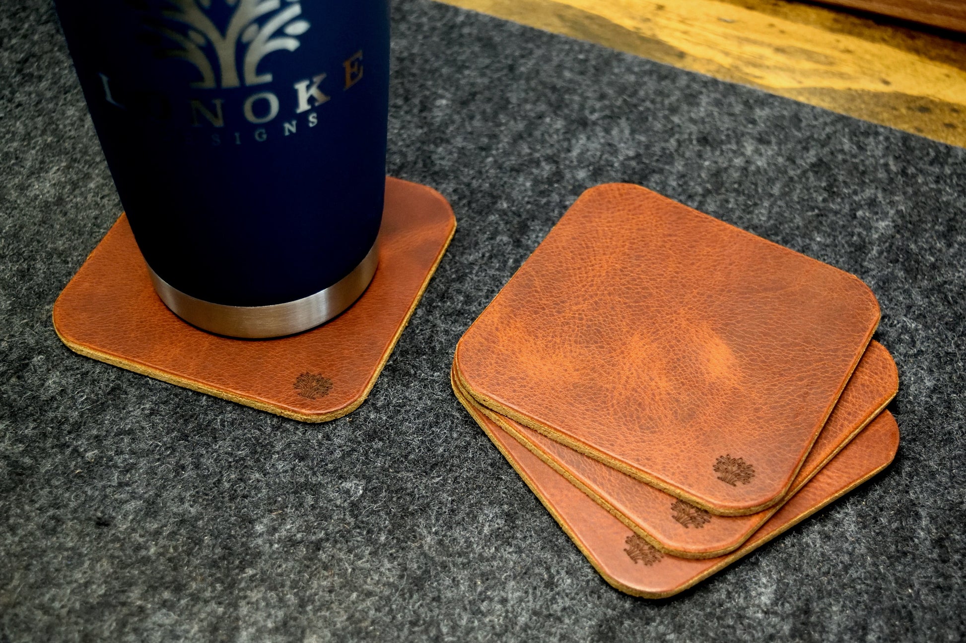 Set of Lonoke Designs brown leather coasters with a blue tumbler on a dark wool surface.