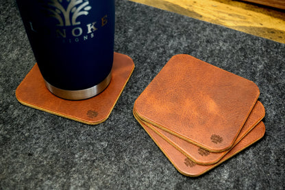 Set of Lonoke Designs brown leather coasters with a blue tumbler on a dark wool surface.