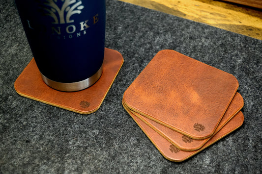 Set of Lonoke Designs brown leather coasters with a blue tumbler on a dark wool surface.