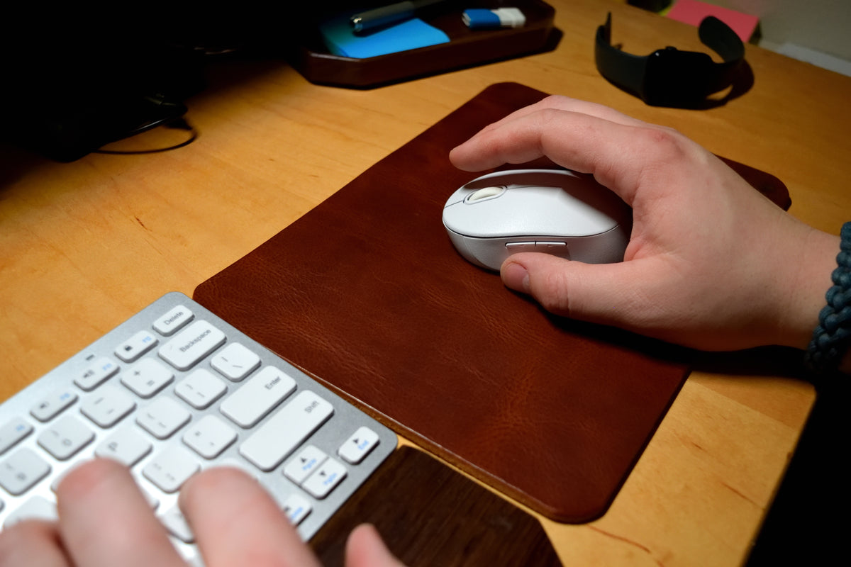 Hand using a wireless mouse on a desk with a keyboard and Lonoke Designs brown leather mouse pad.