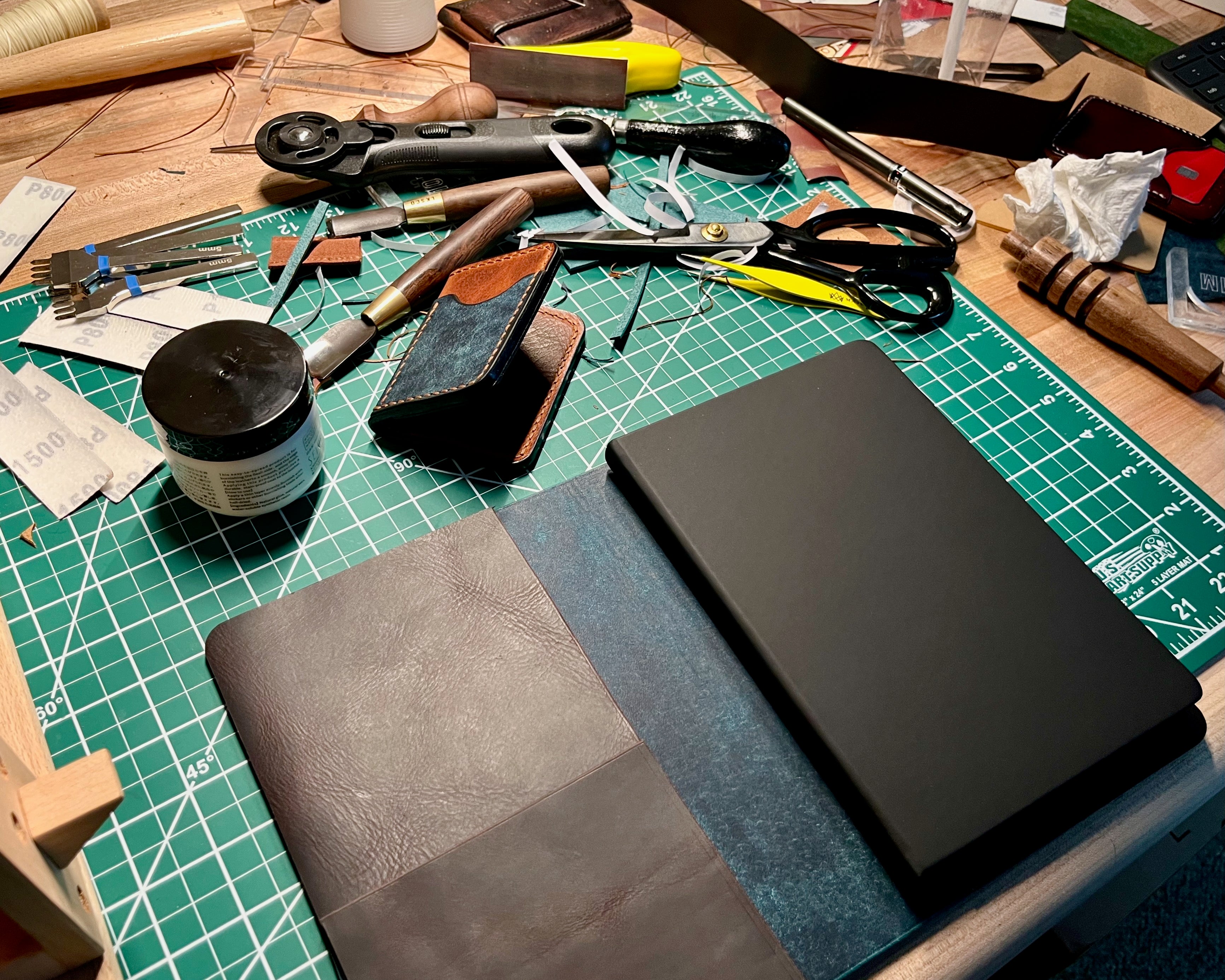 Workbench with leather crafting tools and materials on a green cutting mat.