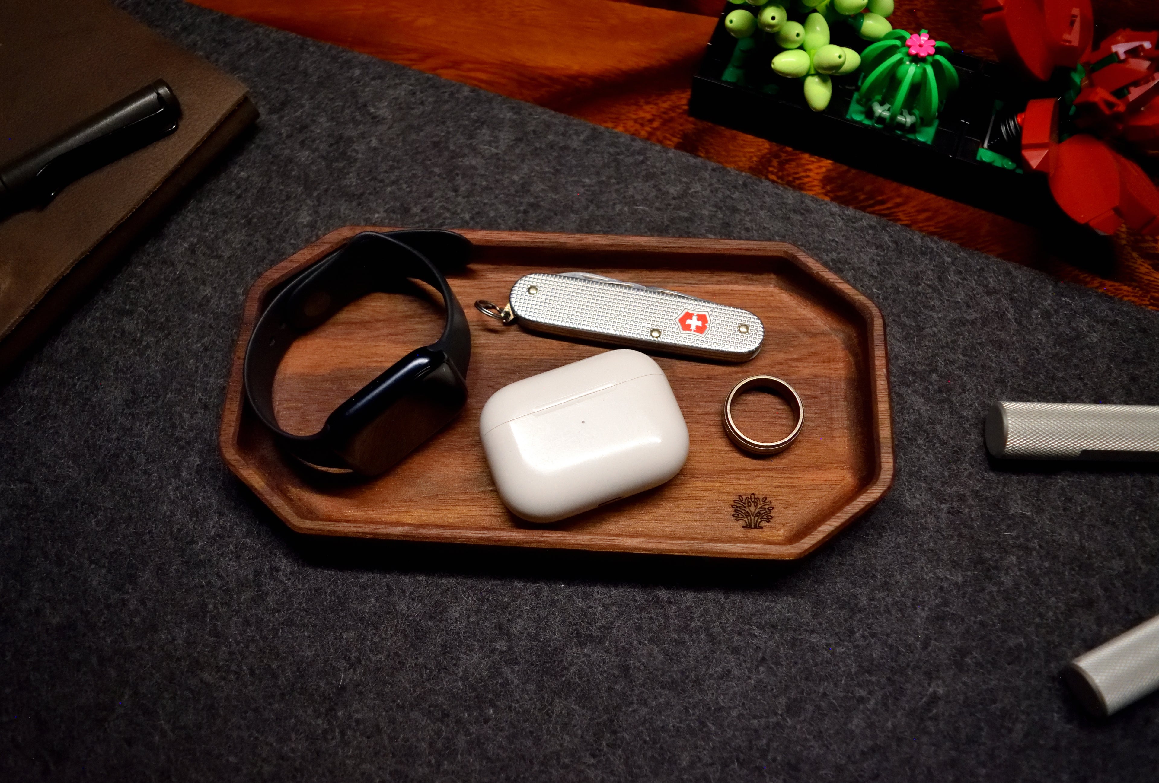 A walnut wood organizer tray displayed on a desk with various items such as a Apple Air Pods, a Swiss Army tool, titanium ring and other desk items arranged on and around it.