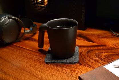 A square coaster made of wool felt and cork, displayed on a wooden surface with a black cup on top of it.