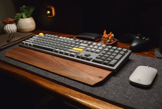 A walnut hardwood wrist rest placed on a wool desk pad with a keyboard and mouse.
