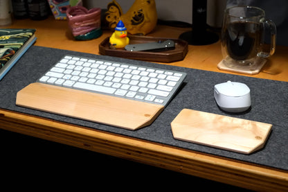 A wireless keyboard and mouse with Lonoke Designs Maple wooden wrist rests are arranged on a dark gray desk mat on a wooden desk, with a mug and other desk items in the background.