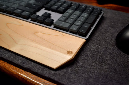 A mechanical keyboard with a Lonoke Designs Maple hardwood wrist rest on a dark wool desk pad.