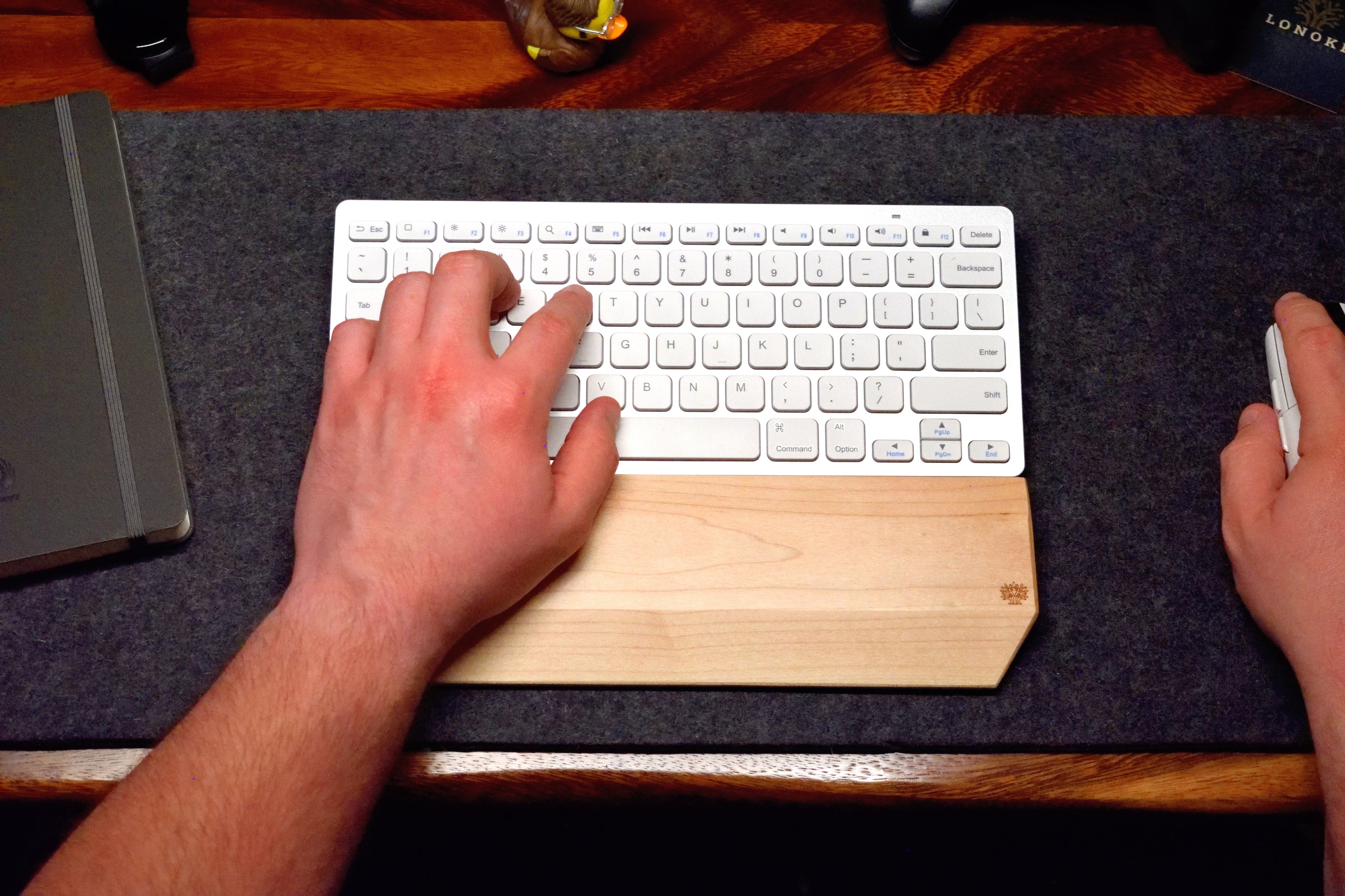 A person typing on a white keyboard, with a Lonoke Designs Maple hardwood wrist rest on a dark wool desk pad.