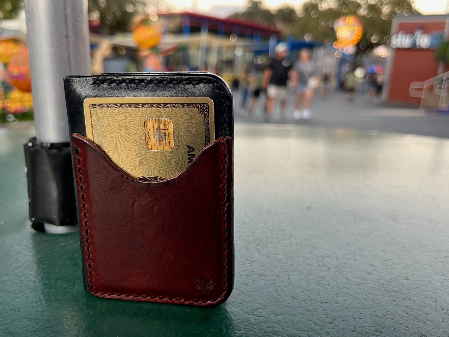 Brown leather Lonoke Designs wallet with a gold card on a blurred amusement park background.