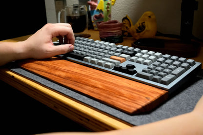 A close-up of a hand using a mechanical keyboard with a Lonoke Designs Red Zebrawood wrist rest on a desk in a dimly lit room.