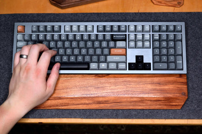 A hand resting on a Lonoke Designs Red Zebrawood wrist rest in front of a silver mechanical keyboard. All resting on a gray wool felt desk pad on a wooden desk.