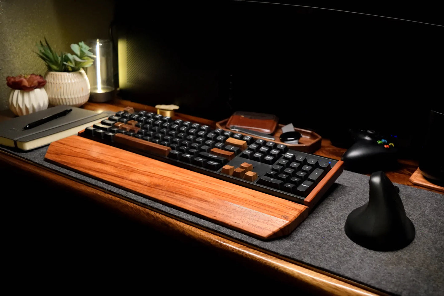 A mechanical keyboard with a prominent Lonoke Designs Red Zebrawood wrist rest and black keycaps, styled on a dark desk wool felt desk pad alongside a game controller, notebook, and plants.