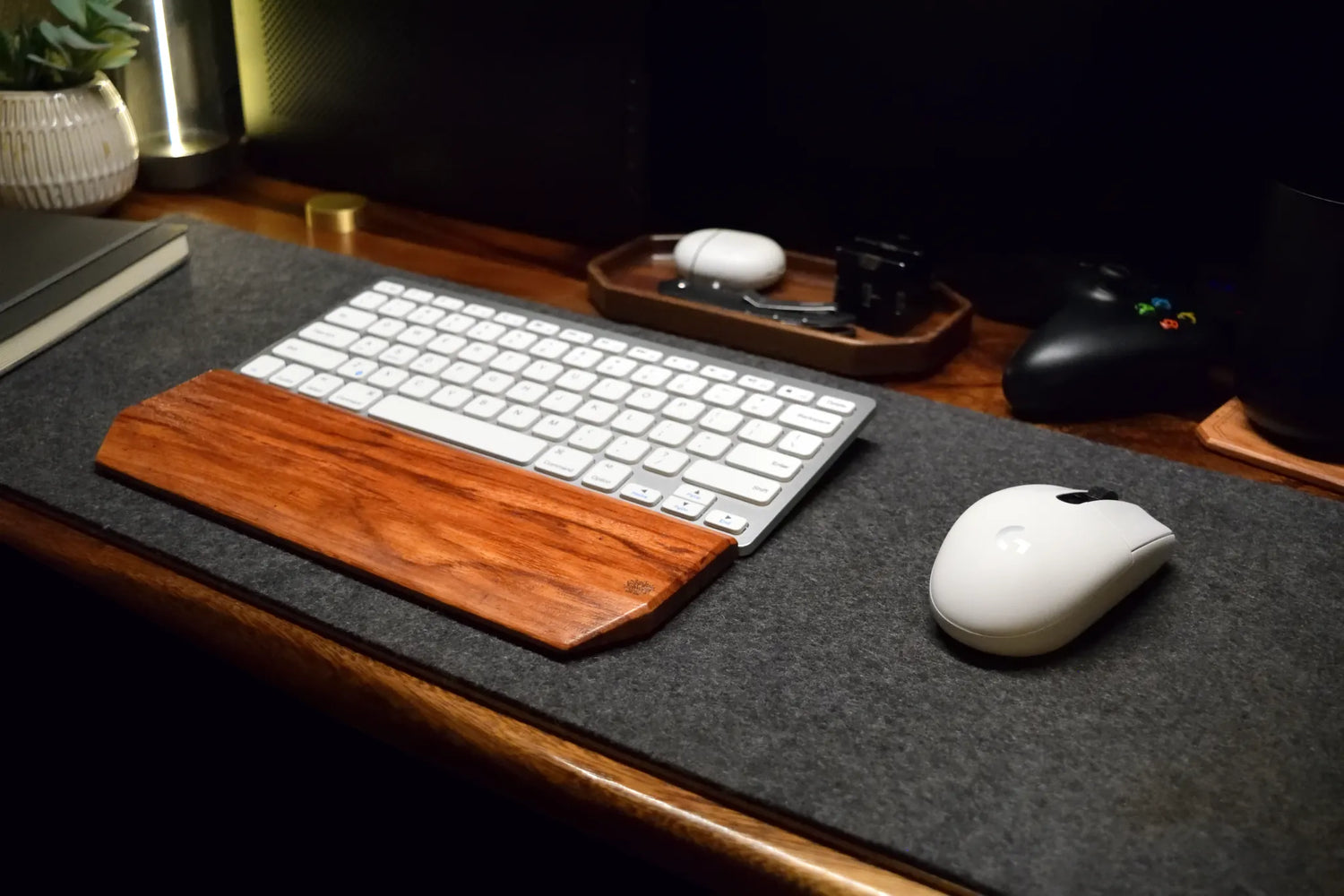 A close-up of a desk setup featuring a white wireless keyboard and mouse resting on a dark gray felt desk mat with a prominent Lonoke Designs Red Zebrawood wrist rest. The scene is set on a wooden desk with warm ambient lighting, showing a game controller and other desk accessories in the background.