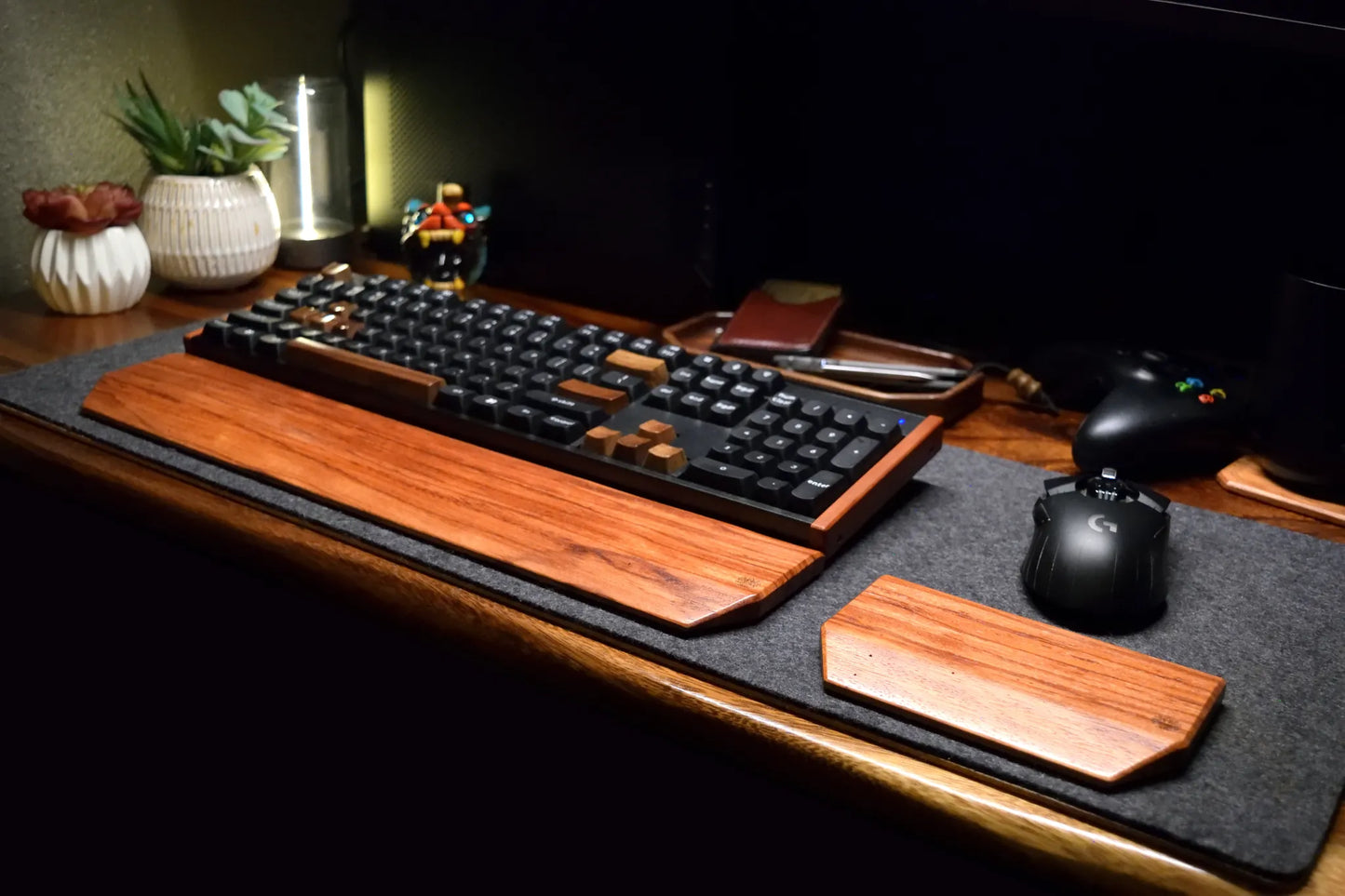 A styled desk setup featuring a black mechanical keyboard and a black gaming mouse resting on a dark gray wool felt desk mat with a Lonoke Designs Red Zebrawood exotic hardwood wrist rest and a matching mouse wrist rest, set against a dark background.