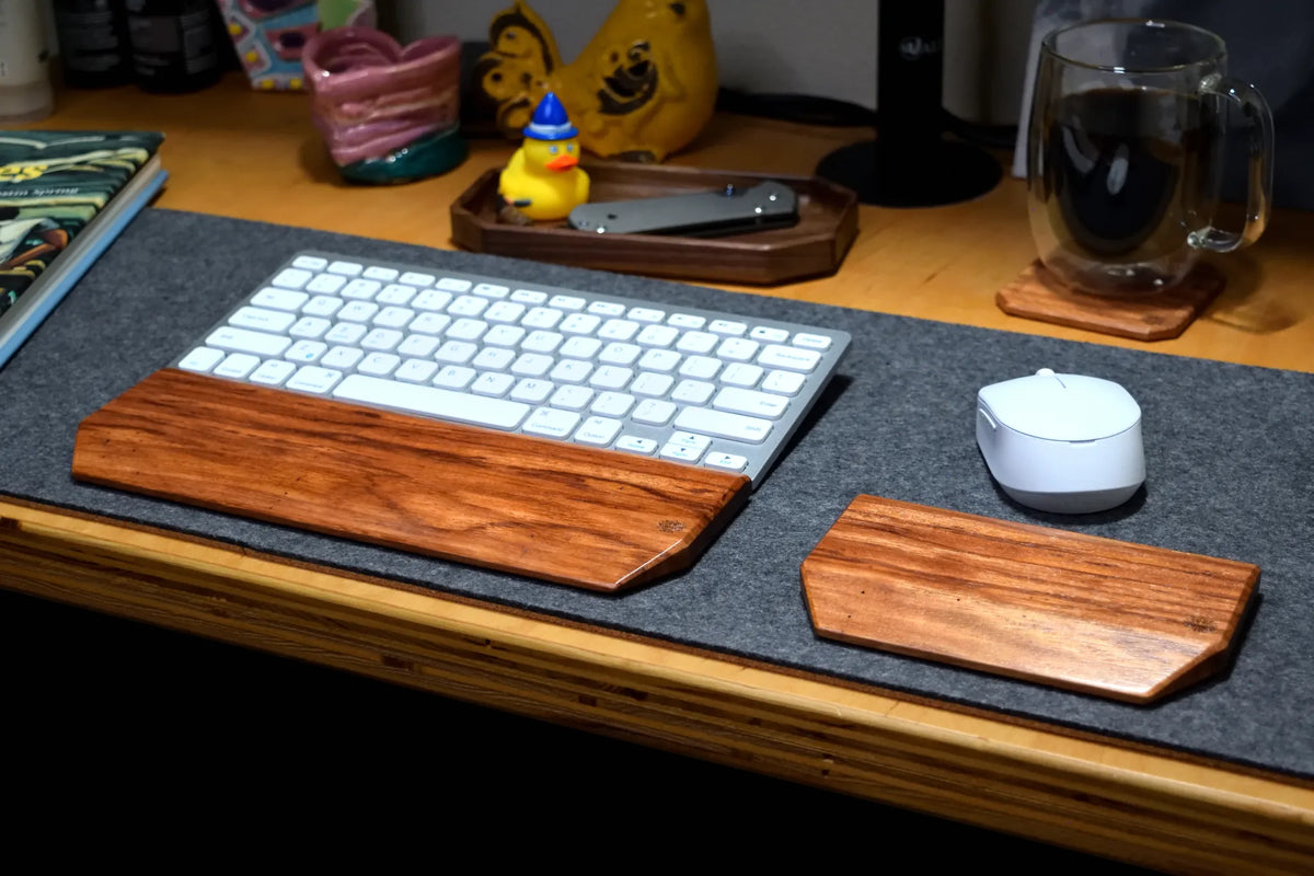 A Lonoke Designs Red Zebrawood keyboard wrist rest and matching mouse rest set arranged on a dark gray desk mat on a wooden desk, alongside a white wireless keyboard and mouse.