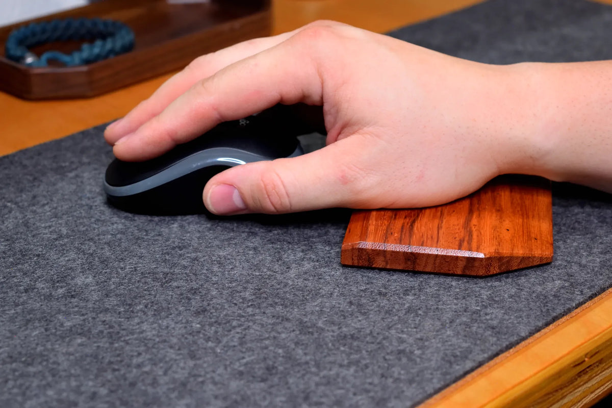 A close-up of a hand resting on a Lonoke Designs Red Zebrawood hardwood mouse wrist rest, using a black-and-gray computer mouse on a gray wool felt and cork desk pad.