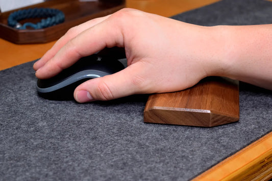 A close-up of a hand resting on a Lonoke Designs walnut hardwood mouse wrist rest, with a black-and-gray computer mouse on a gray wool felt and cork desk pad.