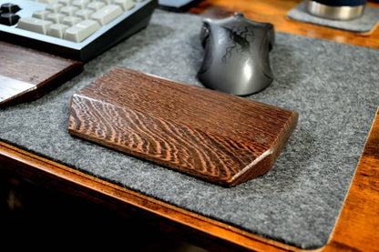 A Lonoke Designs Wenge Mouse Wrist Rest with a prominent wood grain pattern, resting on a gray wool felt desk mat on a wooden desk next to a keyboard and mouse.