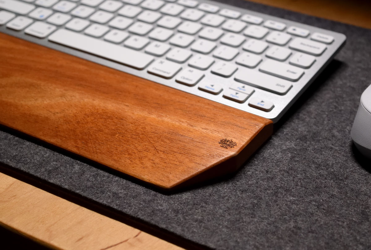A silver-and-white keyboard with a Lonoke Designs Mahogany wood keyboard wrist rest and a white mouse on a charcoal-gray wool felt desk pad. Shown upclose to highlight the flowing wood grain.