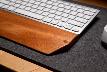 A silver-and-white keyboard with a Lonoke Designs Mahogany wood keyboard wrist rest and a white mouse on a charcoal-gray wool felt desk pad. Shown upclose to highlight the flowing wood grain.
