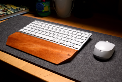 A silver-and-white keyboard with a Lonoke Designs Mahogany wood keyboard wrist rest and a white mouse on a charcoal-gray wool felt desk pad.