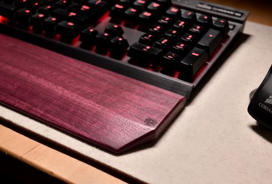 A mechanical gaming keyboard with red backlighting, paired with a Lonoke Designs Purpleheart exotic wood keyboard wrist rest. Shown up close to highlight the woods natural flowing grain.