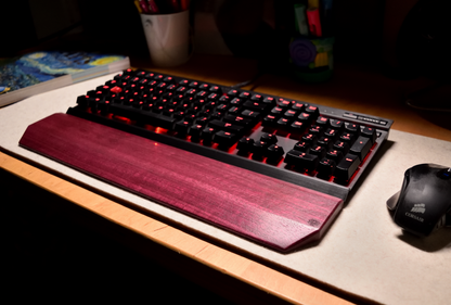 A mechanical gaming keyboard with red backlighting, paired with a Lonoke Designs Purpleheart exotic wood keyboard wrist rest. Shown on a desk next to a mouse in a dimly lit room setting.