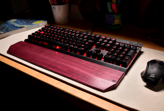 A mechanical gaming keyboard with red backlighting, paired with a Lonoke Designs Purpleheart exotic wood keyboard wrist rest. Shown on a desk next to a mouse in a dimly lit room setting.