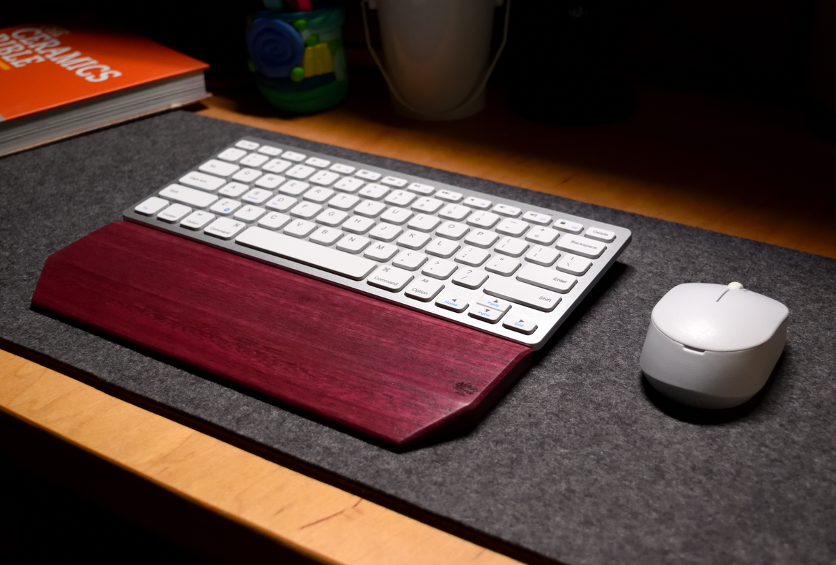 A wireless keyboard paired with a Lonoke Designs Purpleheart exotic wood keyboard wrist rest. Shown on a charcoal wool felt desk pad next to a mouse in a dimly lit room setting.