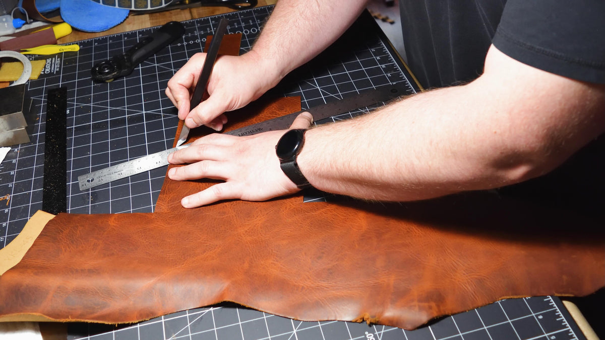 A person's hands using a metal ruler and knife to cut a large piece of brown leather on a black gridded cutting mat at the Lonoke Designs workshop.
