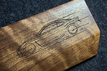 A close-up of a Lonoke Designs walnut mechanical keyboard wrist rest with a laser-engraved sketch of a sports car on its side, resting on a dark textured felt surface.